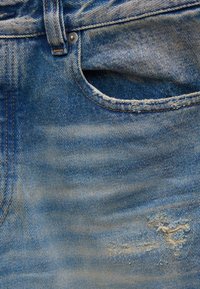 Κοντινή λήψη μπλε τζιν παντελονιού με μπροστινή τσέπη, θηλιά ζώνης, μεταλλικό πρεσαριστό καρφί και μικρό σκισμένο σημάδι κοντά στο κάτω δεξί μέρος.