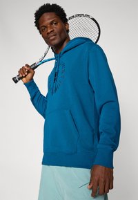 Blå bomulds hoodie med et rundt logo på brystet, kængurulomme og ribbede manchetter. Modellen holder et tennisketsjer over skulderen.