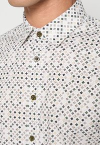 Korte mouwen overhemd met een witte basis, voorzien van een geometrisch patroon van kleine, gekleurde vierkanten in groen, beige en grijs. Voorzien van knopen aan de voorkant.