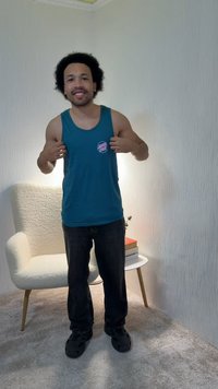 Hombre sonriendo, vestido con camiseta sin mangas azul y pantalones negros, de pie en el interior cerca de un sillón blanco y una mesa pequeña con libros y una planta.