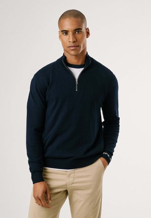 Marine blauwe zip-up sweater met lange mouwen, ribbelmanchetten en -zoom, zachte textuur en een logo-accent op de mouw. Draag over een wit T-shirt.