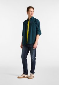 Teal overhemd met knopen over een felgeel t-shirt, gecombineerd met donkere spijkerbroeken en beige sneakers met bruine accenten. Casual ontwerp.