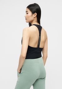 Γυναίκα με μακριά μαύρα μαλλιά που φοράει μαύρο τοπ με τιράντες racerback με κείμενο στις τιράντες και ανοιχτοπράσινο παντελόνι με τα χέρια στις τσέπες.