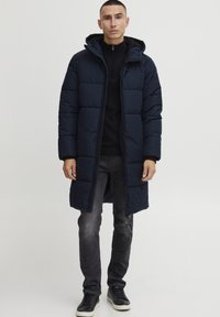 Námořnická puffa bunda s kapucí, která má prošívaný design, dlouhé rukávy, zipový přední uzávěr a postranní kapsy, nošena přes černý top a šedé kalhoty.