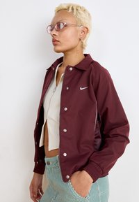 Casaco de nylon cor bordô com botões de pressão e punhos elásticos. Apresenta um logo branco da Nike e painéis laterais, usado sobre uma blusa branca canelada e calças listradas.