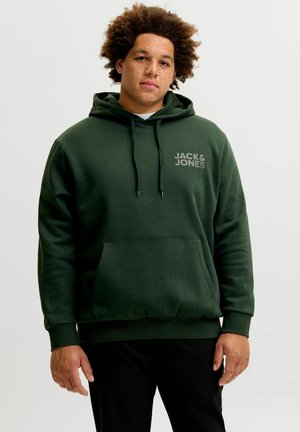 Jeune homme aux cheveux bouclés portant un sweat à capuche vert foncé avec le logo « Jack & Jones », debout devant un fond clair uni.
