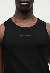 Κοντινό πλάνο ενός ατόμου που φοράει μαύρο ριμπ μπλουζάκι με το κείμενο "KARL LAGERFELD" τυπωμένο σε μικρά, κεντραρισμένα, ανάγλυφα μαύρα γράμματα στο στήθος.