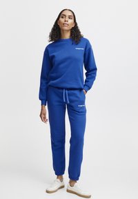 Blå sweatshirt og joggingbukser i blødt stof, med ribbede manchetter og elastisk talje, parret med hvide sneakers. Logo på brystet.