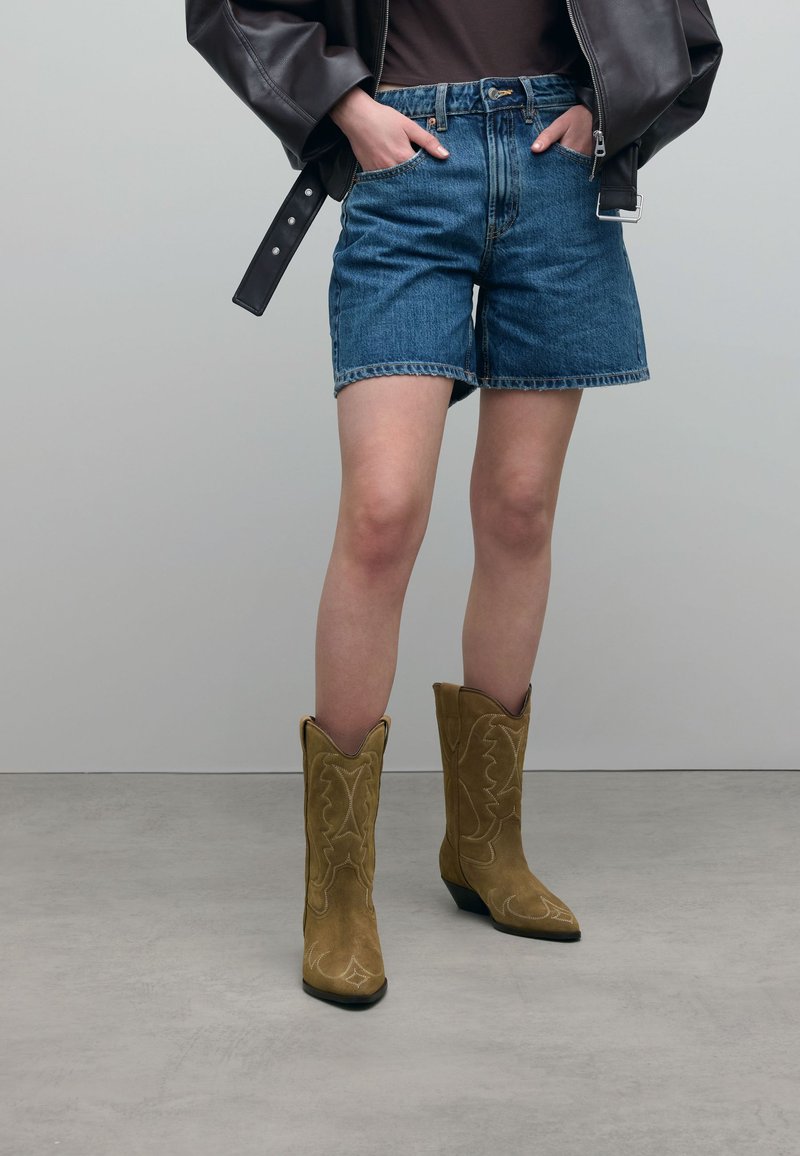 Tan suede cowboy boots with pointed toes and decorative stitching, paired with denim shorts and a dark jacket, against a neutral background.