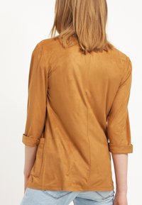 Brun mockakavaj med trekvartslånga ärmar, uppvikt ärm och en avslappnad passform. Har en slät yta och sidofickor.