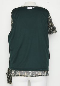 Haut sans manches vert foncé superposé à un chemisier à manches transparentes à fleurs avec poignets froncés, vu de dos sur un mannequin blanc.