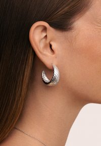 Pendientes de aro de plata con un patrón texturizado similar a escamas, presentando una forma curva y un acabado pulido, mostrados en la oreja de un modelo.