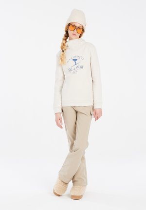 Beige fleece trui met hoge kraag en bedrukte tekst, gecombineerd met beige skibroek en tan fuzzy laarzen. Model draagt een gebreide muts en een bril.