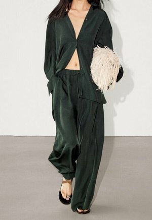 Femme portant une chemise ample en satin vert foncé et un pantalon assorti, tenant une grande pochette crème à franges, marchant pieds nus sur un sol gris.