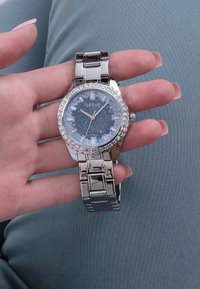 Srebrny zegarek ze stali nierdzewnej z niebieską błyszczącą tarczą, przezroczystymi kamieniami wokół pierścienia i srebrnymi znacznikami godzin.