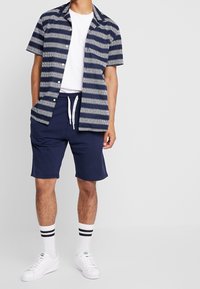 Homme portant une chemise ouverte rayée marine, un t-shirt blanc, un short marine, des chaussettes blanches avec des rayures noires et des baskets blanches, se tenant contre un fond uni.