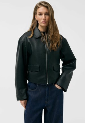 Kobieta z długimi, jasnobrązowymi włosami, ubrana w czarną skórzaną kurtkę zapinaną na zamek i ciemnoniebieskie dżinsy, stojąca z jedną ręką w kieszeni.