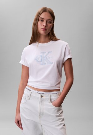 Jeune femme debout, main dans la poche, portant un t-shirt court blanc Calvin Klein et un jean blanc sur fond gris.