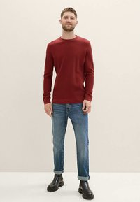 Maglione rosso lavorato a maglia, scollo rotondo, vestibilità slim. Abbinato a jeans in denim azzurro chiaro con risvolti e stivaletti neri lucidi alla caviglia. Sfondo semplice.