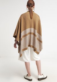 Bruin en beige gebreide poncho met een losse pasvorm, horizontale strepen en een gestructureerde afwerking, gedragen over witte korte broek en schoenen.