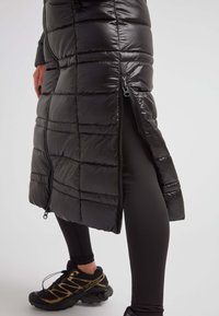 Chaqueta larga acolchada negra con cremallera lateral parcialmente abierta, llevada sobre leggins negros y zapatos de senderismo negros y dorados.