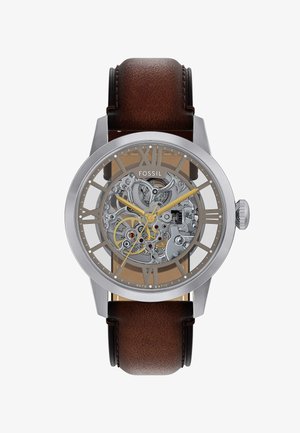 Montre en acier inoxydable argenté avec un cadran squelettique transparent, un bracelet en cuir marron, des accents dorés et de grands index en chiffres romains.
