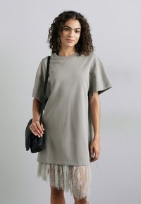 Hallikas üle mõõtmete T-särk pehmest kangast, lühikeste varrukatega ja mitmekihilise pitsservaga. Mudel hoiab käes musta tekstuuriga kotti.