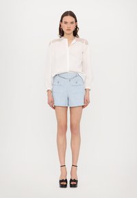Blusa blanca de manga larga con yugo de encaje, combinada con pantalones cortos de tweed azul claro que cuentan con dos bolsillos frontales y detalles metálicos. Zapatos de plataforma negros.