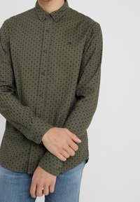 Zielona koszula zapinana na guziki w czarne kropki, wykonana z lekkiego materiału, z długimi rękawami i standardowym kołnierzykiem. Posiada małe logo na klatce piersiowej.