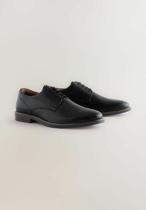 Une paire de chaussures de ville pour hommes en cuir noir avec un système de laçage et des talons en bois empilés sur un fond clair.