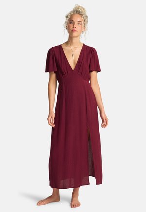 Jeune femme aux cheveux blonds bouclés portant une robe longue rouge foncé à manches courtes avec un col en V et une fente à l'avant, debout pieds nus sur un fond blanc.