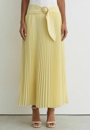 Kvinde iført en lysegul plisseret midi-nederdel med et stofbælte og en guldfarvet cirkulær spænde, kombineret med hvide sandaler med stropper.