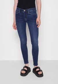 Jeans ajustados azul oscuro con una textura suave, diseño de cinco bolsillos, botón dorado, combinados con sandalias negras gruesas con doble hebilla.