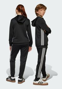 Zwarte sportieve trainingsoutfit met witte strepen aan de zijkanten; hoodie met trekkoorden; bijpassende joggers; sportieve schoenen met contrasterende zolen.