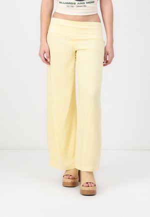 Woman wearing pale yellow wide-leg pants and beige platform open-toe sandals, standing against a plain white background.