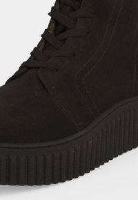 Gros plan d'une botte noire en daim à lacets avec une semelle épaisse à rainures et des coutures visibles sur le côté.