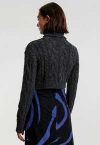 Dunkelgrauer Zopfmuster-Pullover mit hoher Kragen und cropped Design, mit strukturierten Mustern und geripptem Saum, getragen über einem bunten Rock.