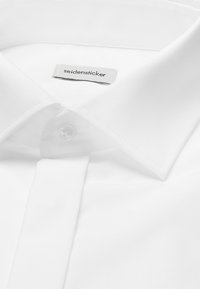 Gros plan sur le col d'une chemise blanche avec un bouton et une étiquette visible portant la mention "seidensticker" à l'intérieur du col.