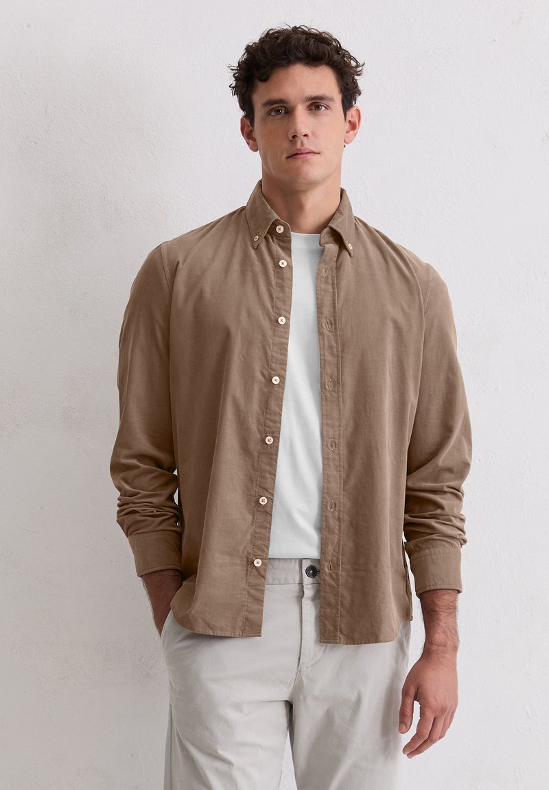 Mężczyzna z kręconymi włosami, ubrany w beżową koszulę z guzikami na białą koszulkę i jasne spodnie, stojący przed gładką białą ścianą.