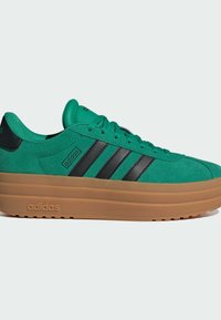 Baskets en suède vert avec des rayures noires, dotées d'une semelle épaisse en gomme et d'un logo brodé sur le côté. Design épuré et surface texturée.