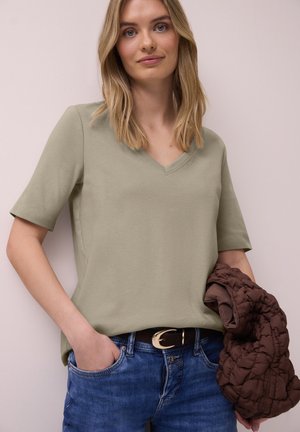 Frau trägt ein hellolivgrünes V-Ausschnitt-T-Shirt mit kurzen Ärmeln und blaue Jeans mit schwarzem Gürtel, hält in der linken Hand eine braune gesteppte Jacke.