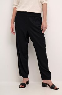 Schwarze, lässig geschnittene Hose aus glattem Stoff mit elastischem Bund. Kombiniert mit schwarzen Riemchensandalen und einem strukturierten cremefarbenen Oberteil.