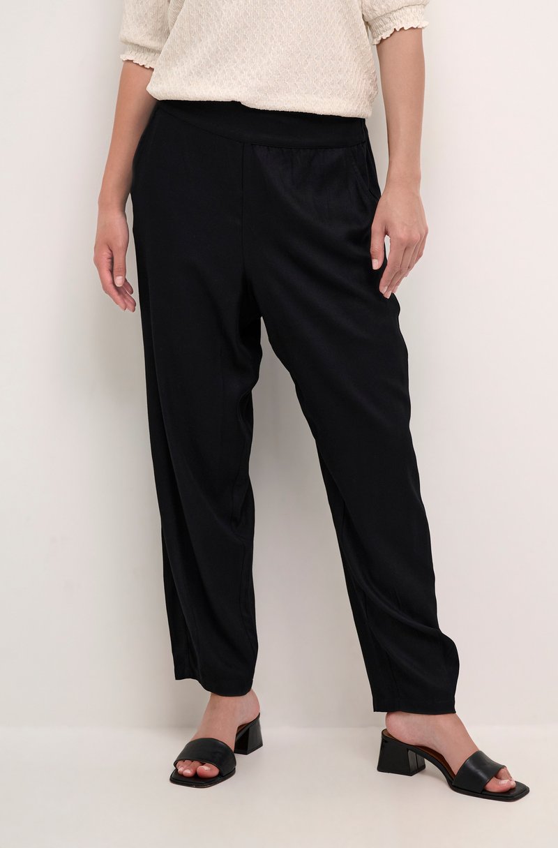 Schwarze, lässig geschnittene Hose aus glattem Stoff mit elastischem Bund. Kombiniert mit schwarzen Riemchensandalen und einem strukturierten cremefarbenen Oberteil.