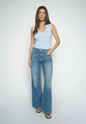 Frau trägt ein ärmelloses Oberteil mit hellblauen und weißen Streifen, hoch taillierte, weit geschnittene Jeans und braune Sandalen, steht vor einem einfarbigen Hintergrund.