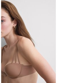 Młoda kobieta o jasnej cerze i brązowych włosach, ubrana w beżowy biustonosz bez ramiączek, widoczna od ramion do połowy tułowia na białym tle.