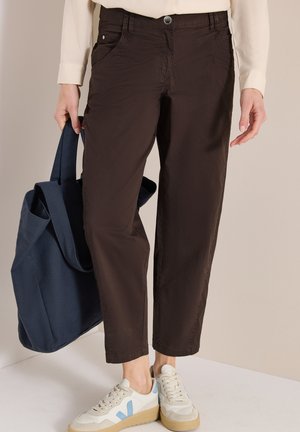 Personne portant un pantalon court marron foncé, une chemise beige, des baskets blanches avec des détails bleus, tenant un sac fourre-tout bleu marine.
