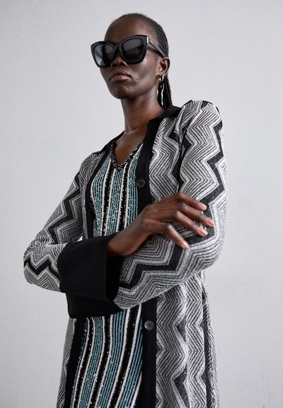 Juodai balta zigzag rašto paltas su juodu apykakle, derinamas su dryžuota, daugiaspalve vidine suknele, dekoruota karoliukais ir blizgučiais.