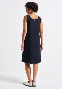 Granatowa sukienka bez rękawów z okrągłym dekoltem, z tylnym detalem w postaci guzika oraz luźnym krojem, zestawiona z białymi i niebieskimi sneakersami.