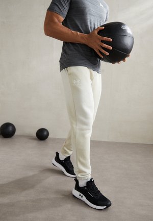 Hellgraue Sporthosen mit schmal zulaufenden Beinen und schwarzer Markenemblematik, kombiniert mit schwarzen Turnschuhen und einem schwarzen Medizinball, in einer Fitnessstudio-Umgebung.