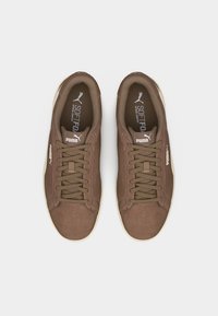 Καφέ σουέντ αθλητικά παπούτσια Puma με μπεζ σόλες και κορδόνια, φαίνονται από πάνω σε λευκό φόντο.